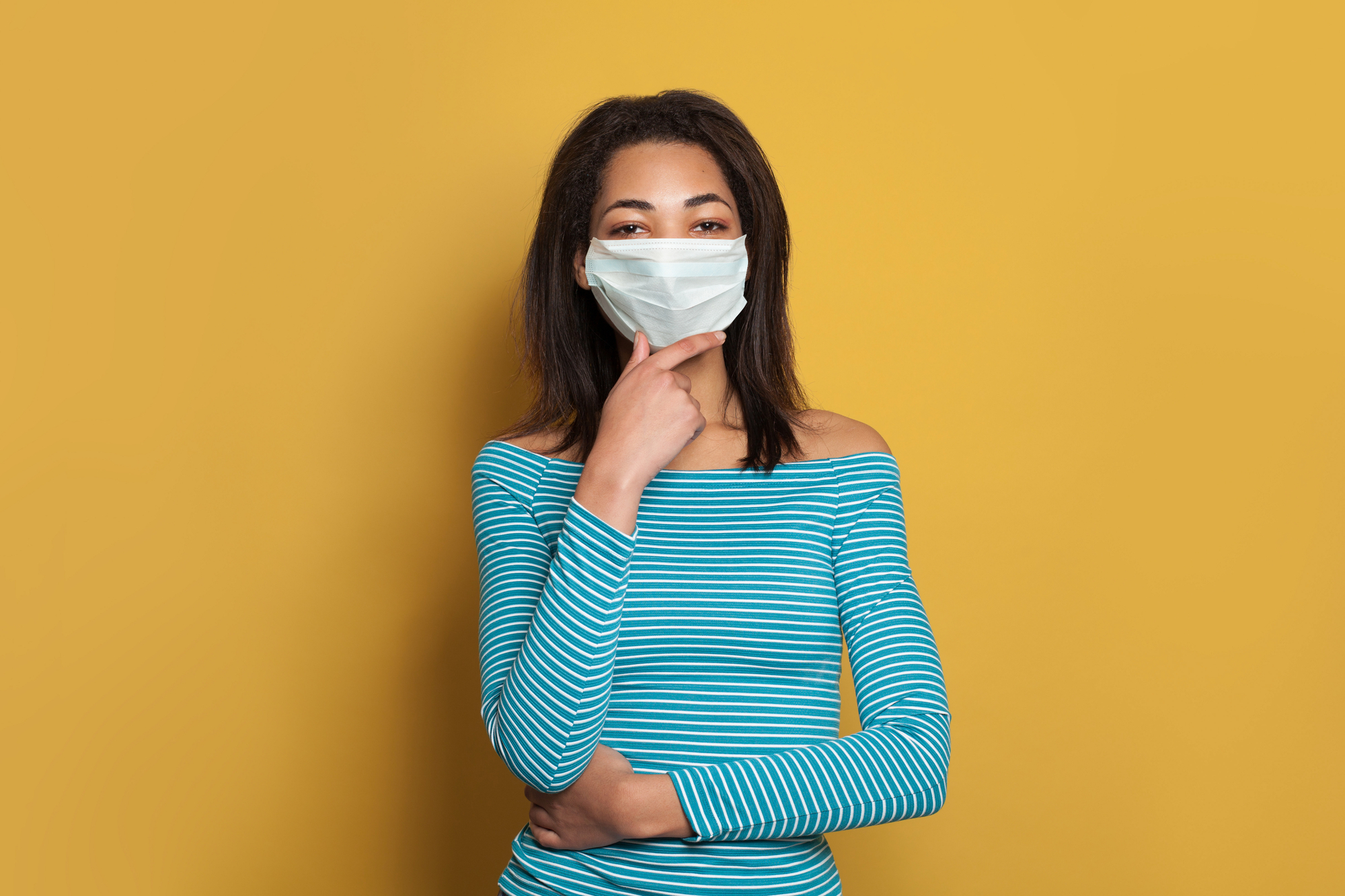 Black woman in medical safety face mask thinking on yellow background