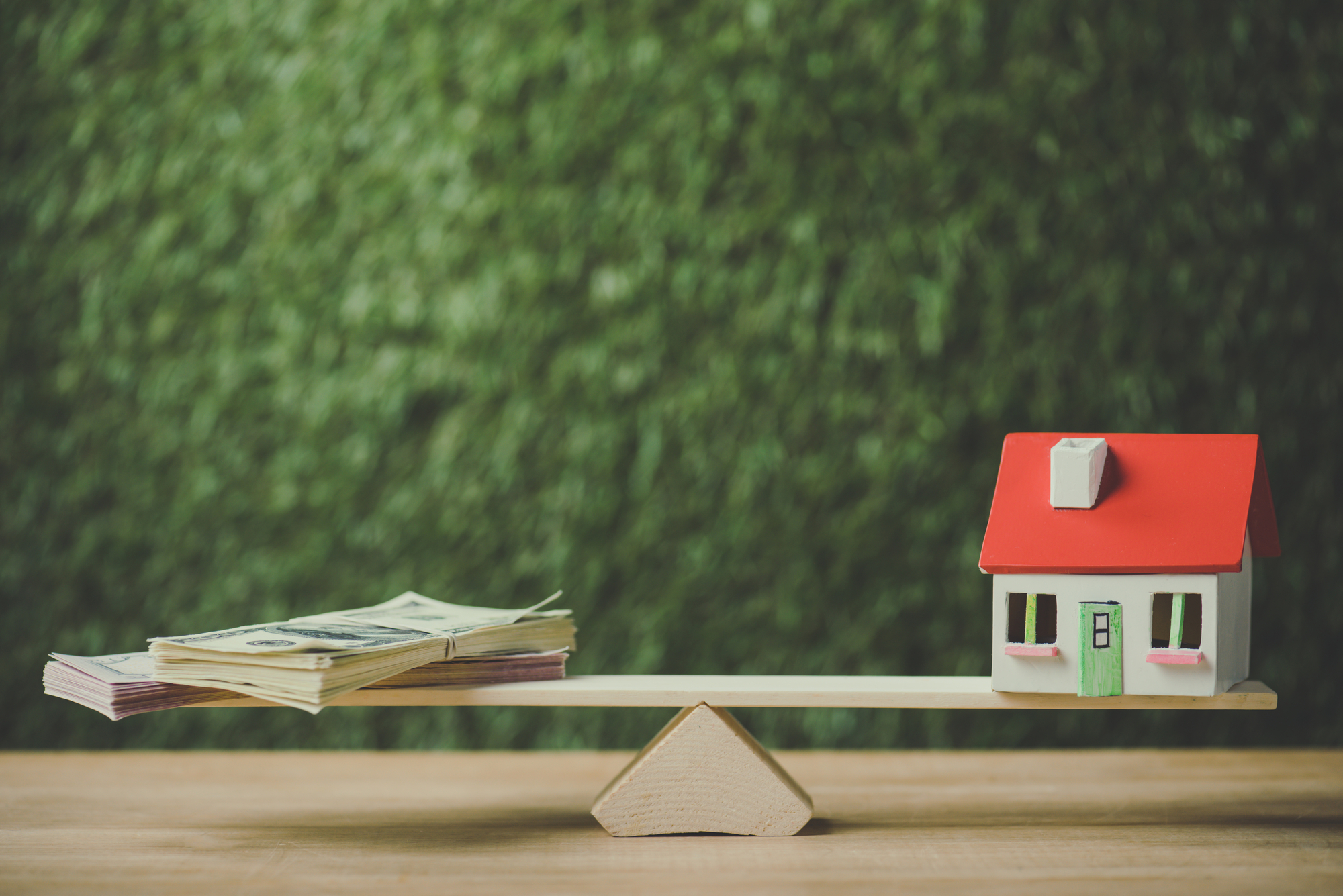 House model and dollar banknotes balancing on seesaw on green background